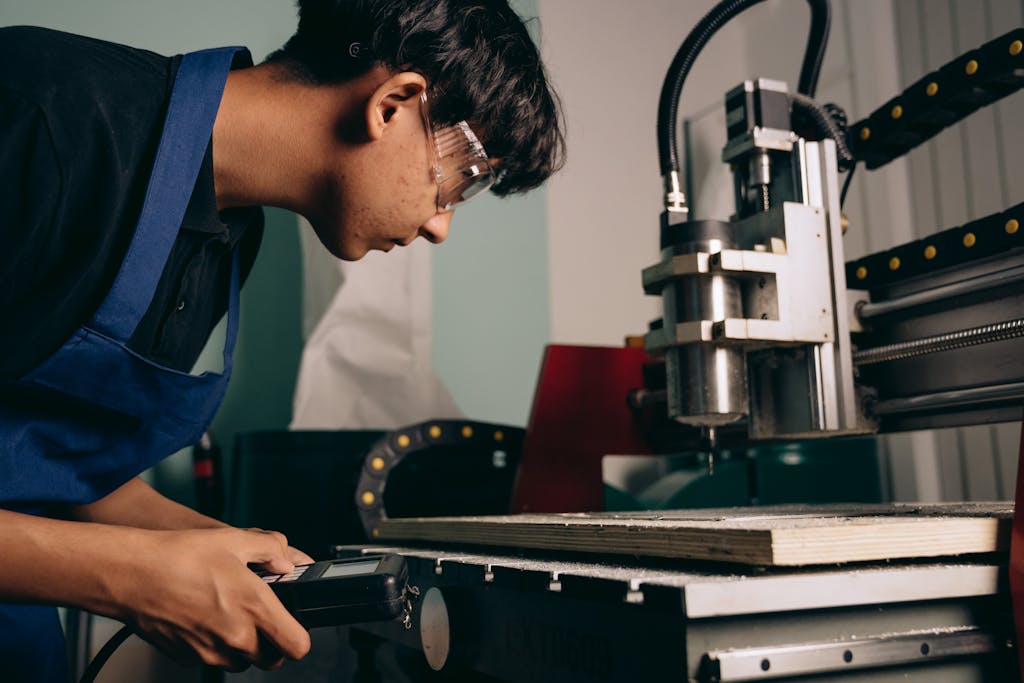 A focused young man wearing safety goggles operates a CNC machine in an industrial workshop.